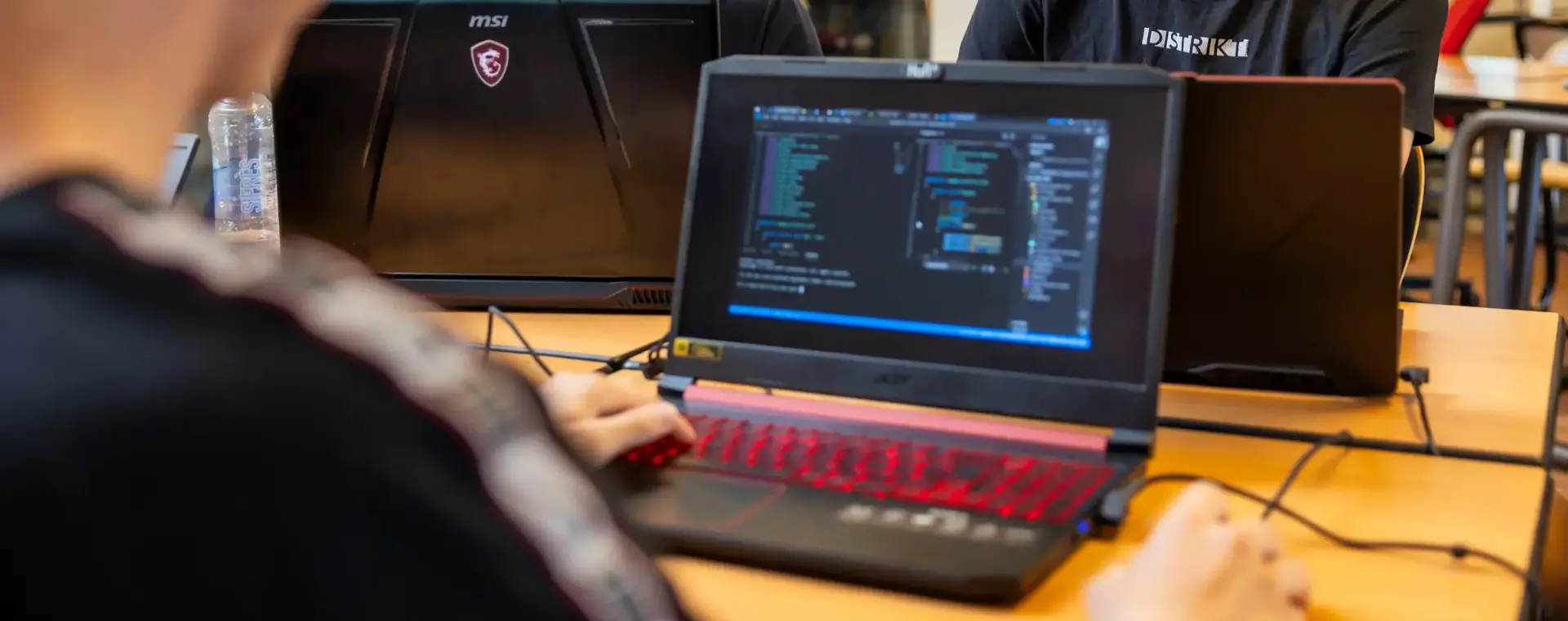 Drie studenten werken samen aan een programmeeropdracht. Ze zitten met hun laptops aan een tafel en overleggen enthousiast over hun code.