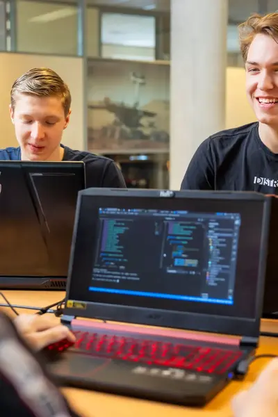 Drie studenten werken samen aan een programmeeropdracht. Ze zitten met hun laptops aan een tafel en overleggen enthousiast over hun code.