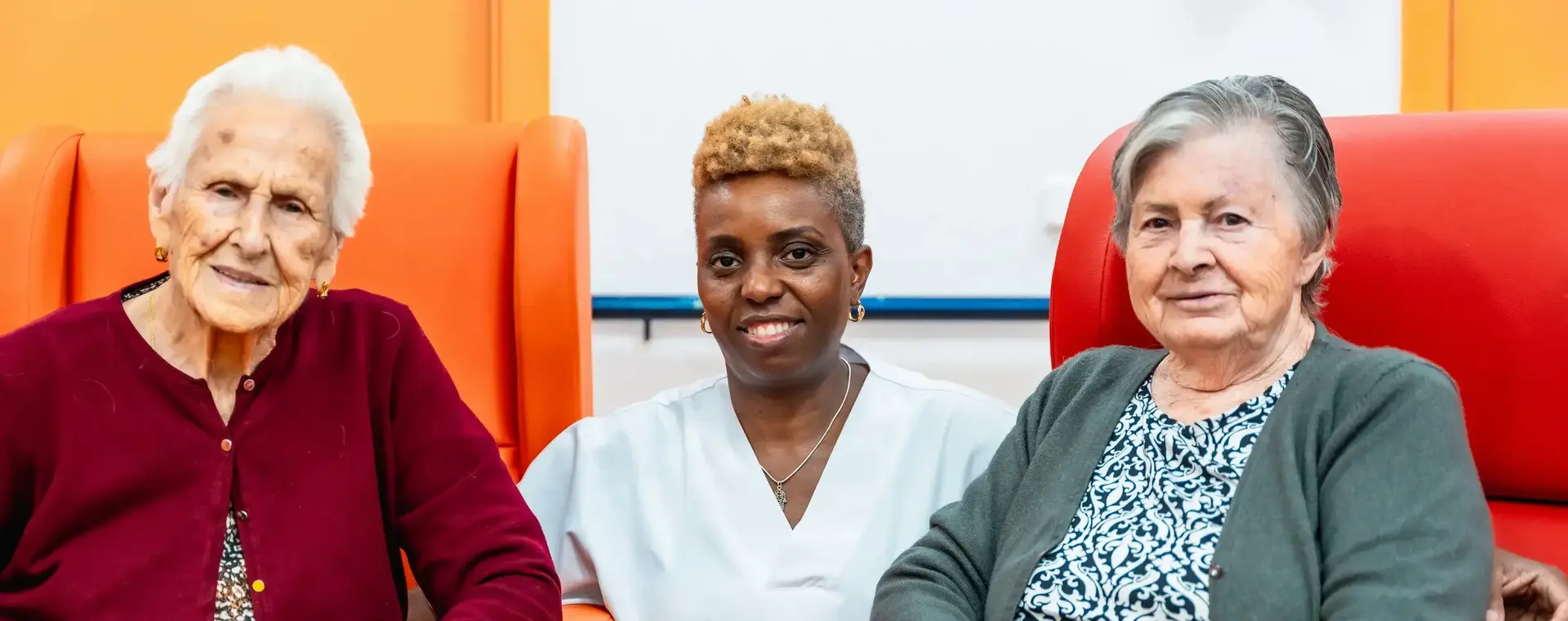 Verpleegster samen met twee bejaarde vrouwen, de bejaarde vrouwen zitten in stoelen en de verpleegster zit gehurkt tussen hen in.