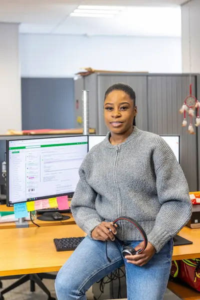 Medewerker zit op een bureau met headset in haar hand en twee computerschermen op de achtergrond.
