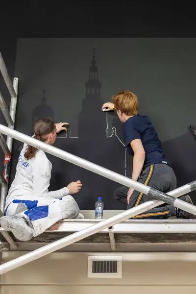 Drie studenten op een bouwstelling zijn een muurschildering aan het maken.