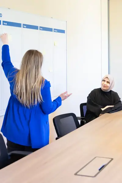 Student schrijft iets op een scrum board terwijl vier andere studenten meedenken tijdens een projectoverleg in een klaslokaal.