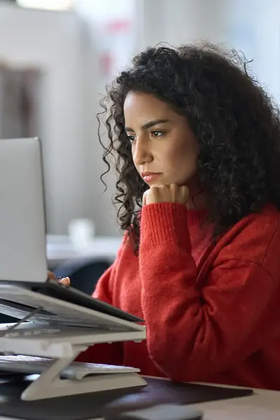 Student werkt geconcentreerd achter een laptop op kantoor. Ze draagt een rode trui en steunt met haar hand onder haar kin terwijl ze naar het scherm kijkt.