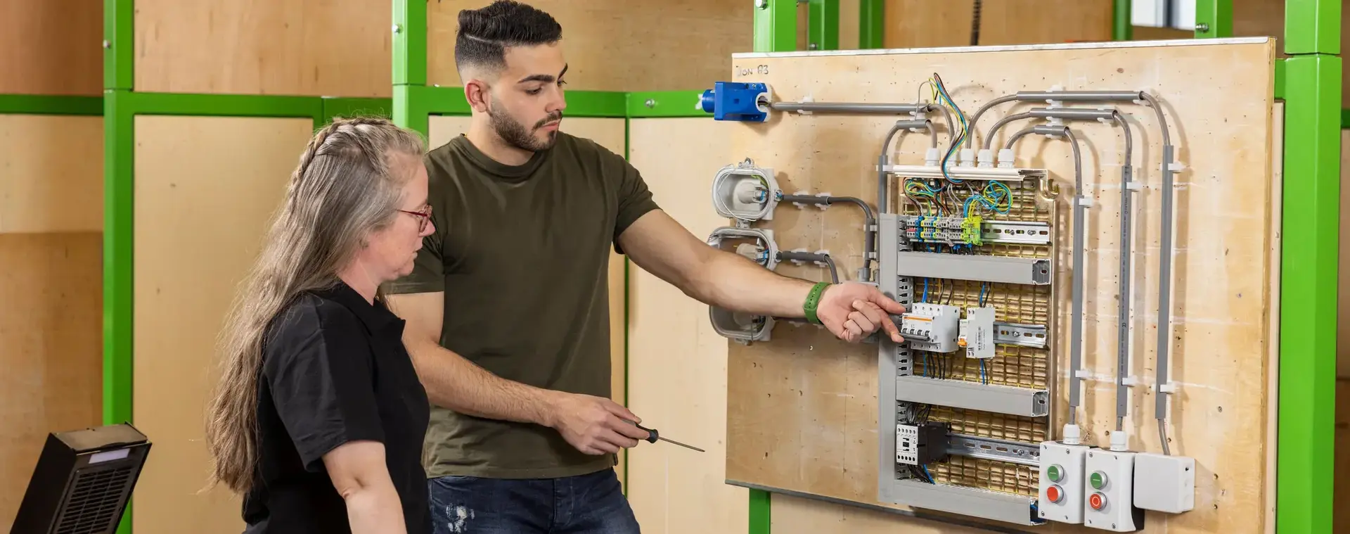 Student legt bedrading uit aan docent bij een oefeninstallatie met stopcontacten, schakelaars en elektriciteitskabels in een lokaal met groene kaders en houten panelen.