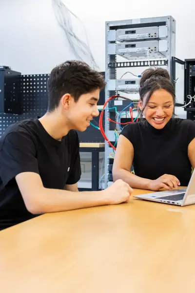 Twee studenten werken samen achter een laptop in een ICT-lokaal. Op de achtergrond staan netwerkapparaten en bekabeling opgesteld.