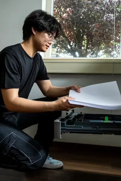Een student in zwarte kleding hurkt voor een grote printer terwijl hij een stapel papier in de lade legt.