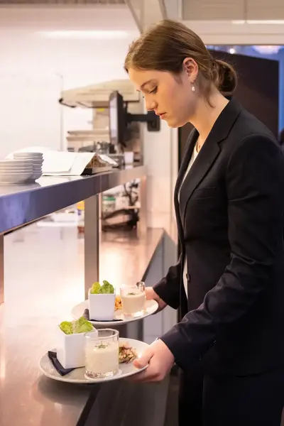 Een jonge vrouw in pak loopt geconcentreerd met twee borden eten de keuken uit. Op de achtergrond zijn collega's in een keuken aan het werk.