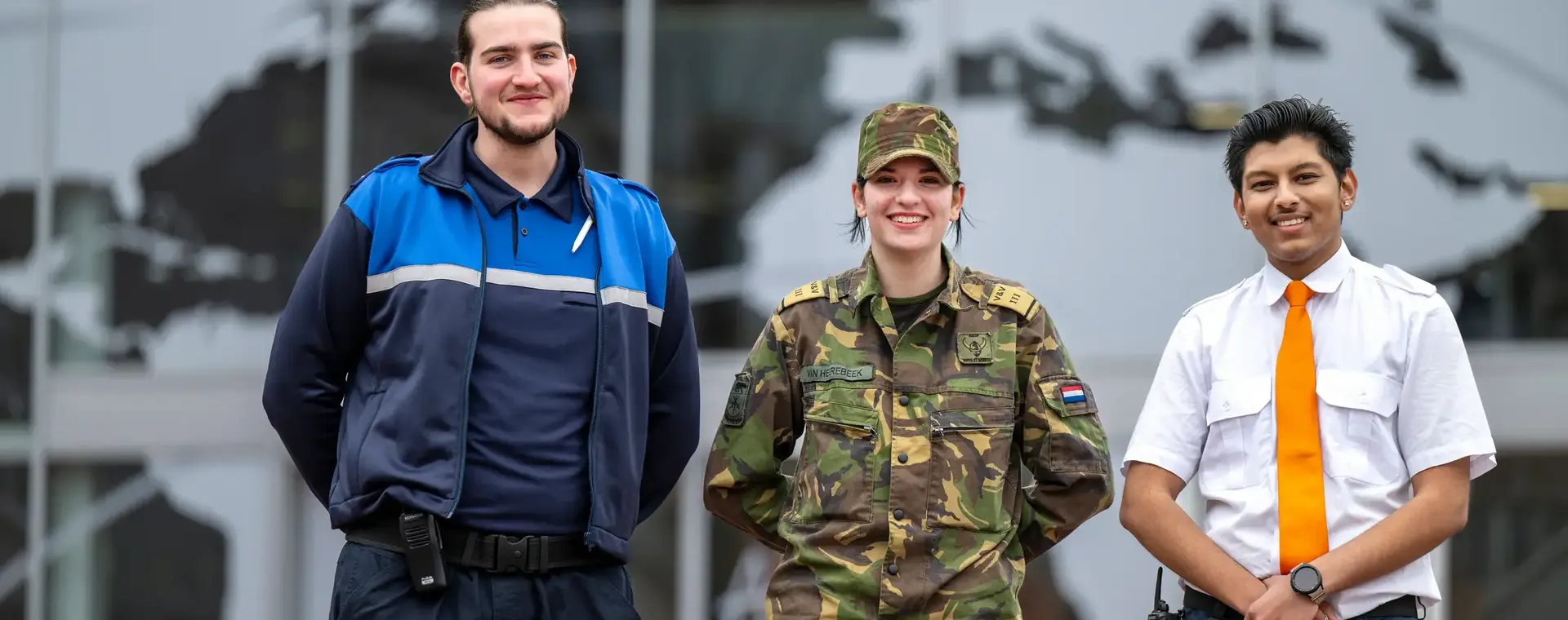 Drie studenten in uniform – beveiliging, defensie en handhaving – poseren trots voor hun schoolgebouw.
