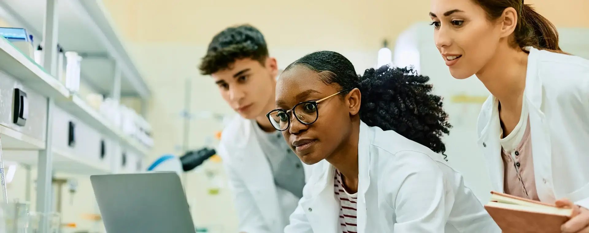 Twee studenten en een docent werken samen aan een onderzoeksopdracht in het lab. Ze analyseren gegevens op een laptop en bespreken hun bevindingen.