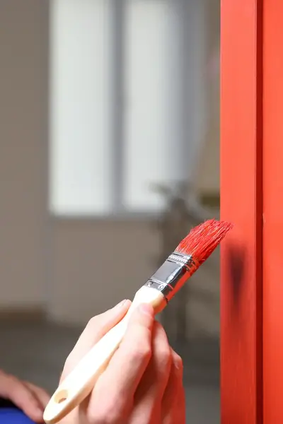 Man met overall aan en glimlach op zijn gezicht, is een kozijn rood aan het schilderen met een kwast.