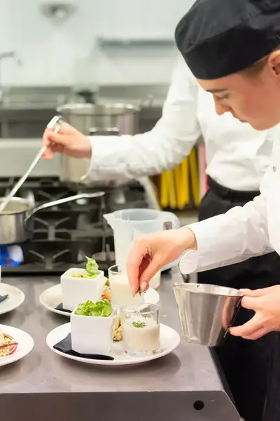 Kok in witte jas en zwarte muts dresseert een bord met salade en mousse in een keuken.