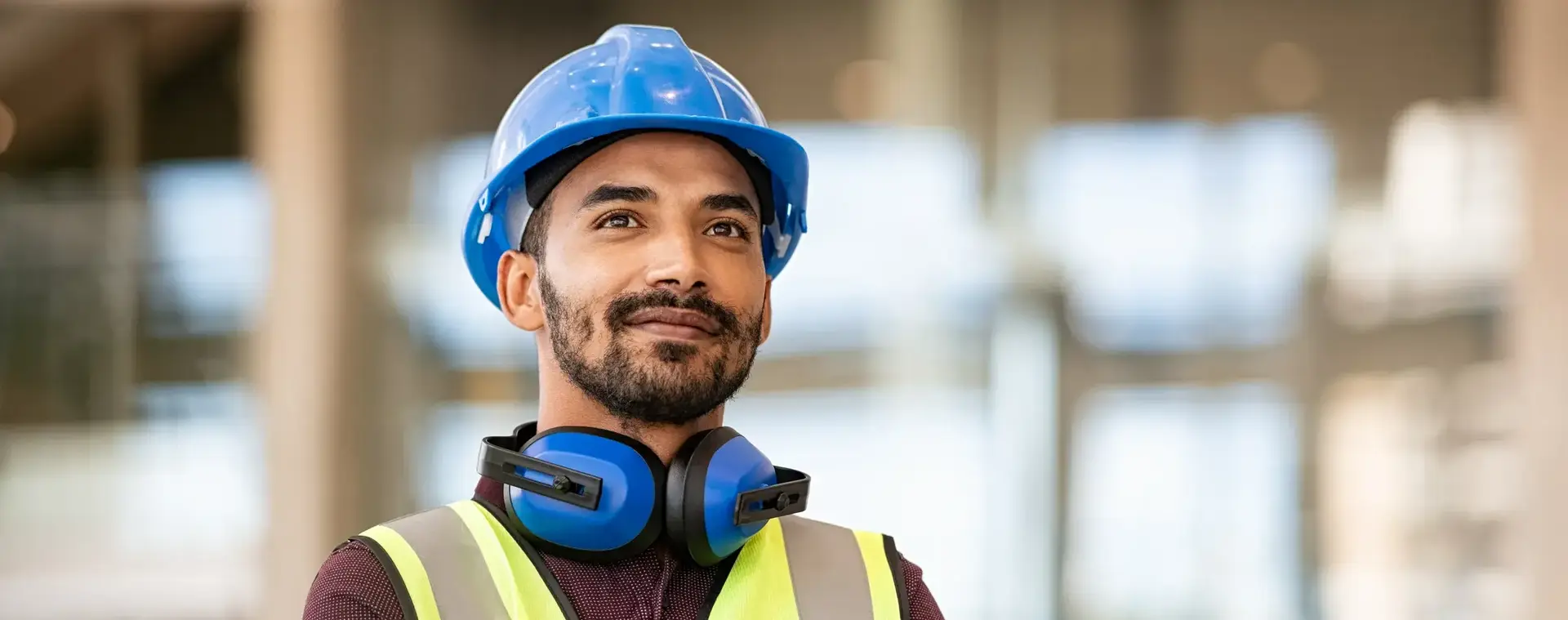 Een man met helm, gehoorbescherming en een geel hesje staat met zijn armen over elkaar en glimlacht een beetje. Op de achtergrond zie je een gebouw.