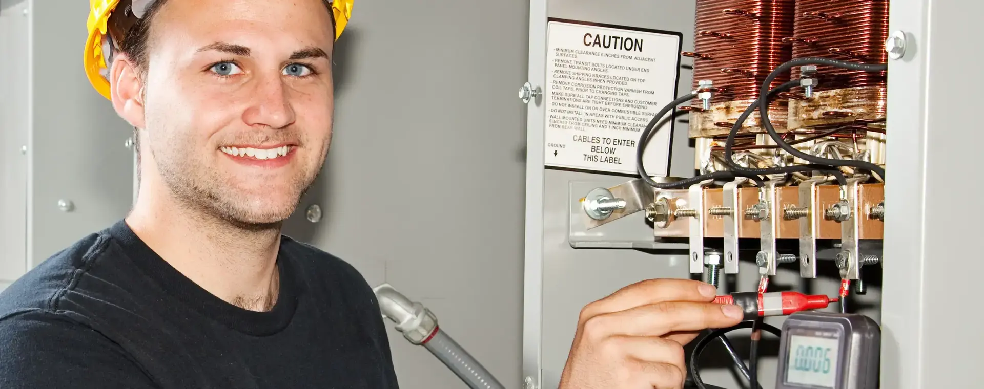 Student met gele helm en meetapparatuur controleert een elektrische installatie in een praktijklokaal. Hij glimlacht naar de camera terwijl hij metingen uitvoert in een verdeelkast.