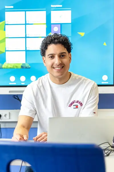 Docent zit achter een laptop in een klaslokaal en kijkt lachend naar de camera.