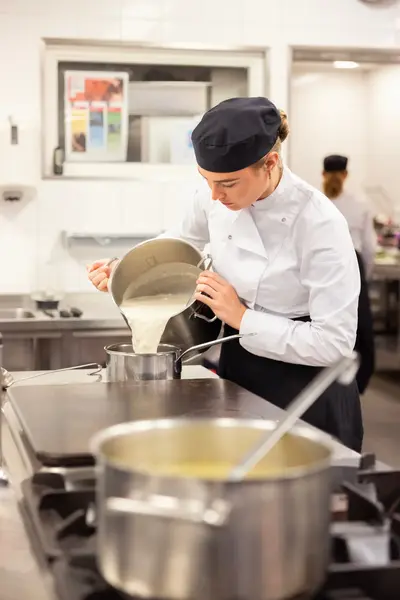 Een kok in witte jas en zwarte pet giet een romige saus in een pan in een professionele keuken.