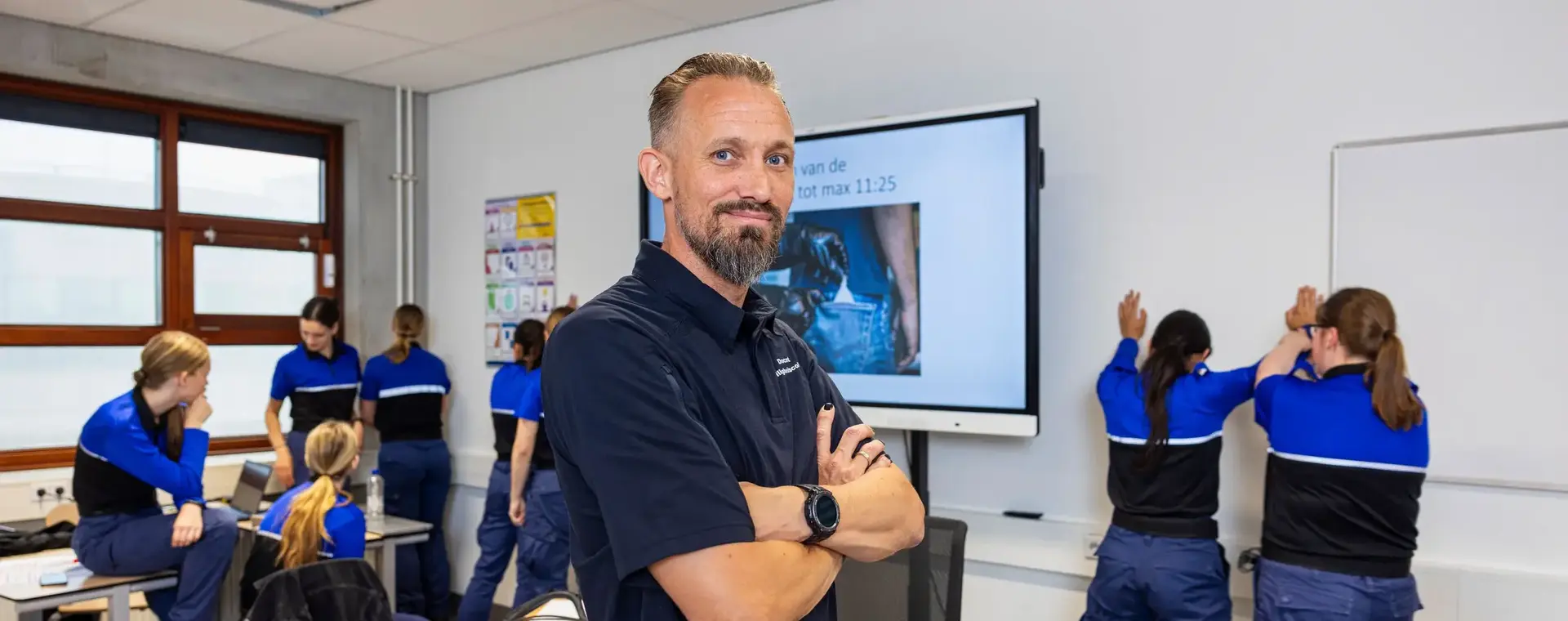Docent veiligheid staat met over elkaar geslagen armen voor de klas. Studenten Veiligheid in uniform zijn op de achtergrond bezig met veiligheidsoefeningen.
