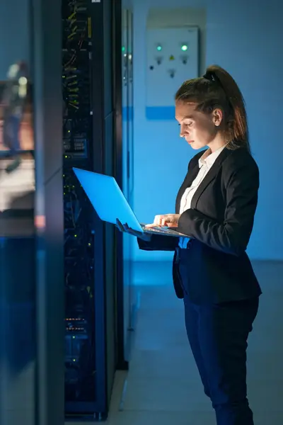 Student werkt met een laptop in een serverruimte. Ze draagt een donker pak en staat tussen verlichte serverkasten terwijl ze gegevens controleert.