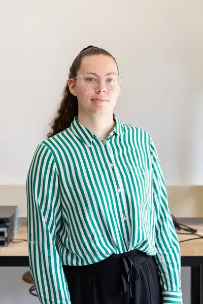 Student poseert in een ICT-lokaal met netwerkapparatuur op de achtergrond. Ze draagt een groen-wit gestreept overhemd en kijkt recht in de camera.