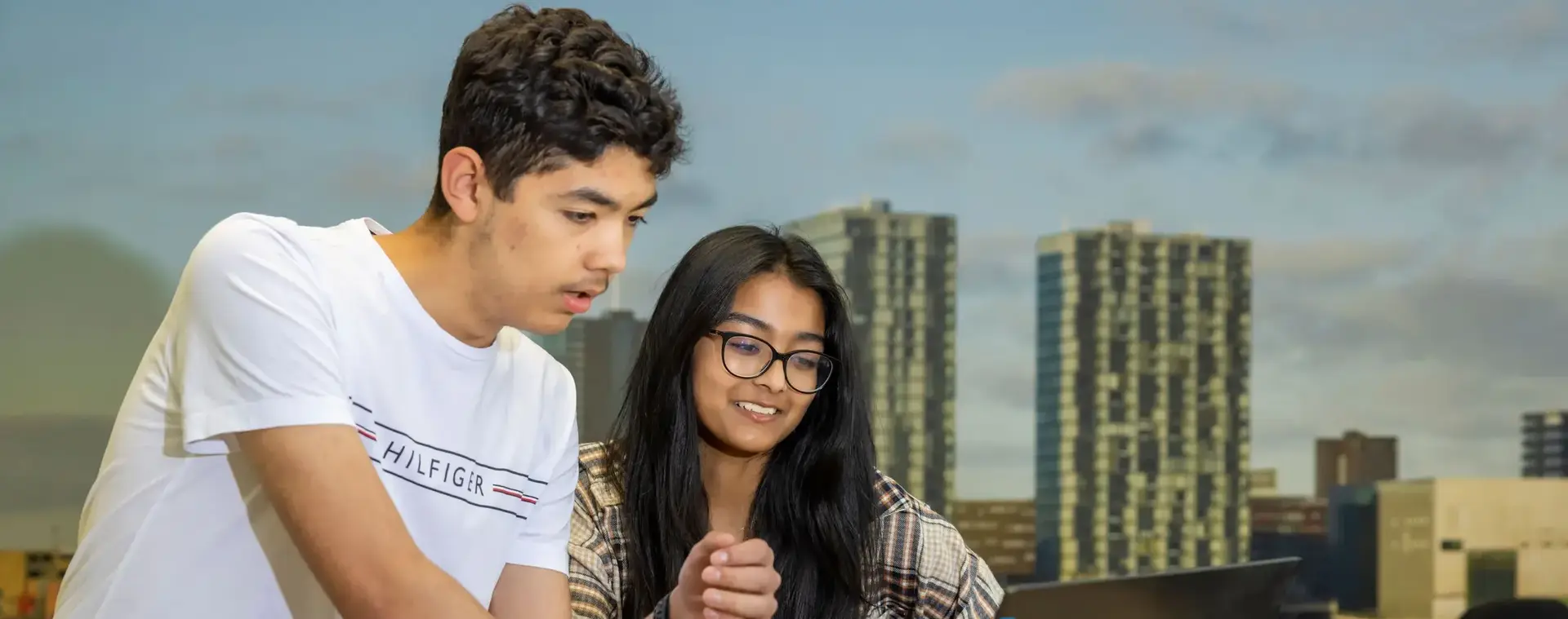 Twee studenten werken samen achter een laptop, waarbij één student iets uitlegt terwijl de ander glimlachend toekijkt, met een skyline van hoge gebouwen en water op de achtergrond.