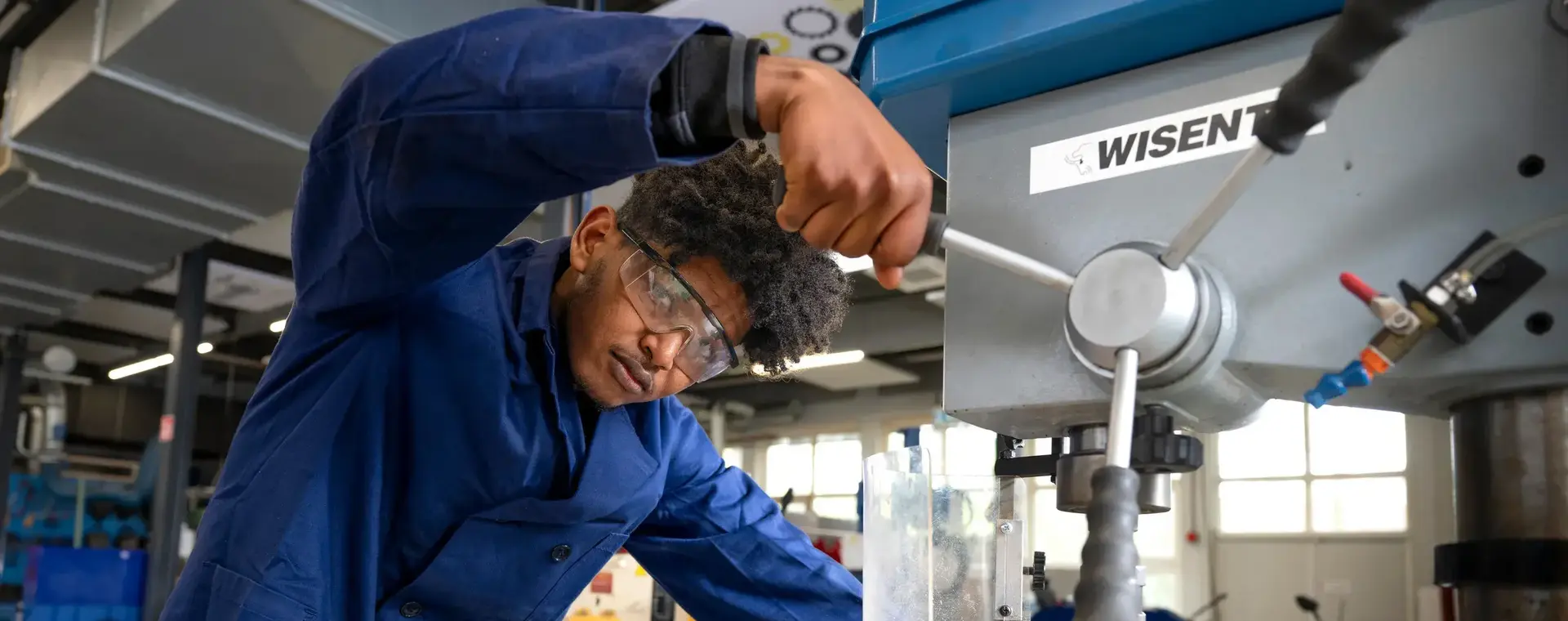 Student met veiligheidsbril en werkjas bedient een grote boormachine in de werkplaats. Hij werkt geconcentreerd en precies aan een houten plaat, met focus op veiligheid.