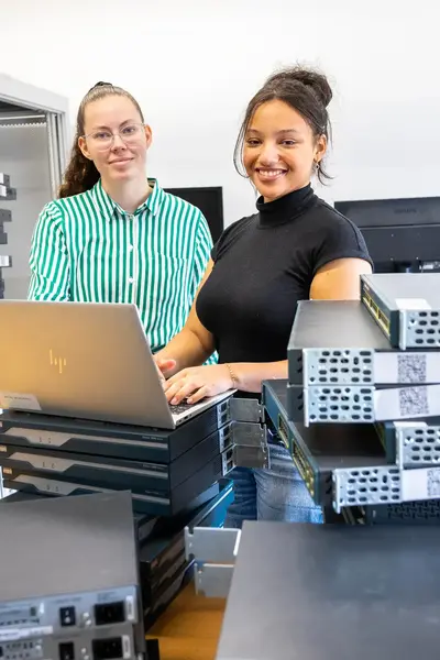Twee studenten werken samen achter een laptop in een ICT-lokaal. Op tafel liggen stapels netwerkapparatuur en op de achtergrond staan serverkasten.