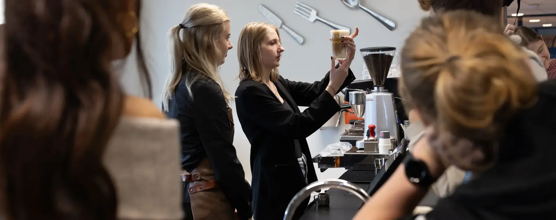 Een docent houdt een glas met koffie omhoog terwijl ze iets uitlegt aan een groep aandachtige studenten in een praktijklokaal met barista-apparatuur.
