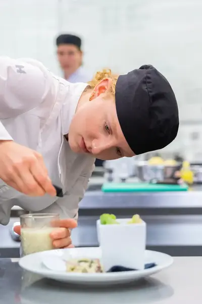 Een kok in witte outfit en zwarte muts schenkt een witte saus in een glas op een bord.