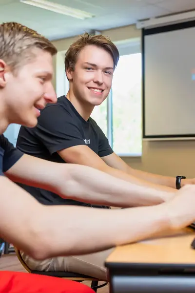 Twee studenten werken samen achter laptops in een computerlokaal. Op de achtergrond staat een projectiescherm met het Microsoft-logo. Eén student kijkt lachend in de camera.
