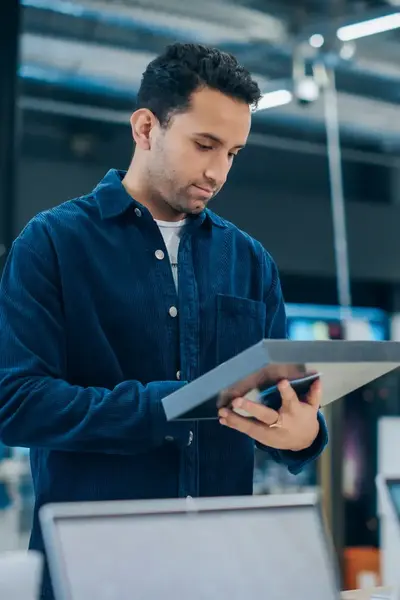 Man bekijkt een tablet in een elektronica winkel. Hij draagt een donkerblauw overhemd en staat tussen uitgestalde laptops en andere apparaten.