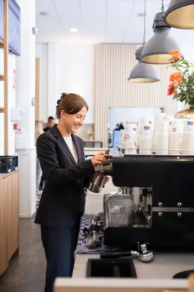 Een jonge vrouw in een donker pak bereidt koffie achter een espressomachine. Ze lacht terwijl ze melk opschuimt.