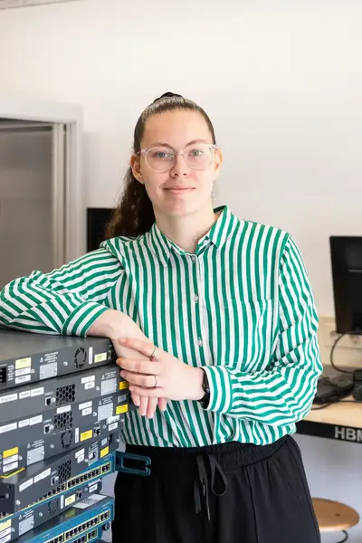 Student poseert in een ICT-lokaal naast een stapel netwerkapparatuur. Op de achtergrond zijn serverkasten en computers te zien.