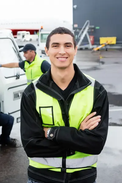 Een luchthavenmedewerker met veiligheidshesje lacht, terwijl zijn collega’s op de achtergrond bij een voertuig zitten.