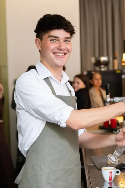 Een jonge man in een schort staat lachend achter de bar en tapt een drankje. Op de achtergrond werken collega's.