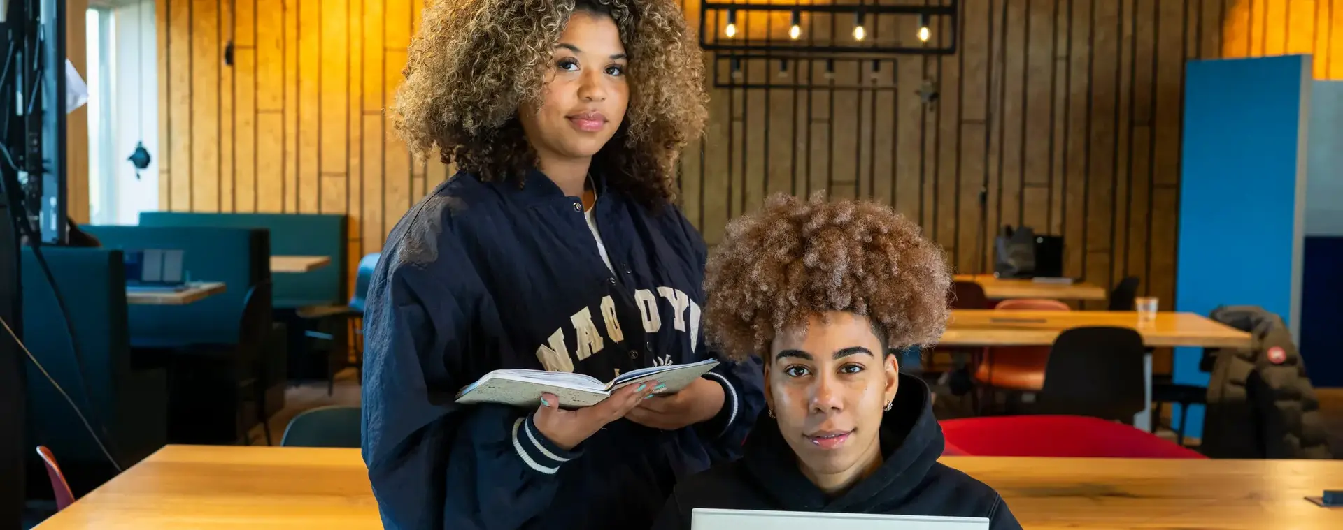 Twee studenten werken samen aan een opdracht in een gezellige leeromgeving. Eén student houdt een laptop vast, terwijl de ander aantekeningen maakt in een notitieboek.