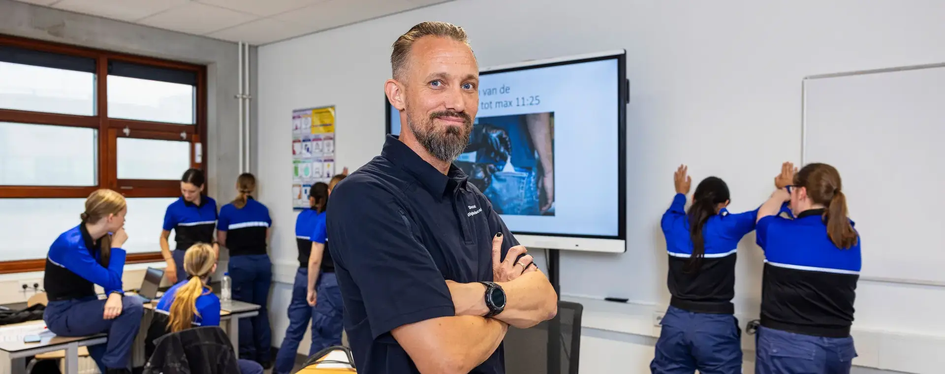 Docent veiligheid staat met over elkaar geslagen armen voor de klas. Studenten Veiligheid in uniform zijn op de achtergrond bezig met veiligheidsoefeningen.