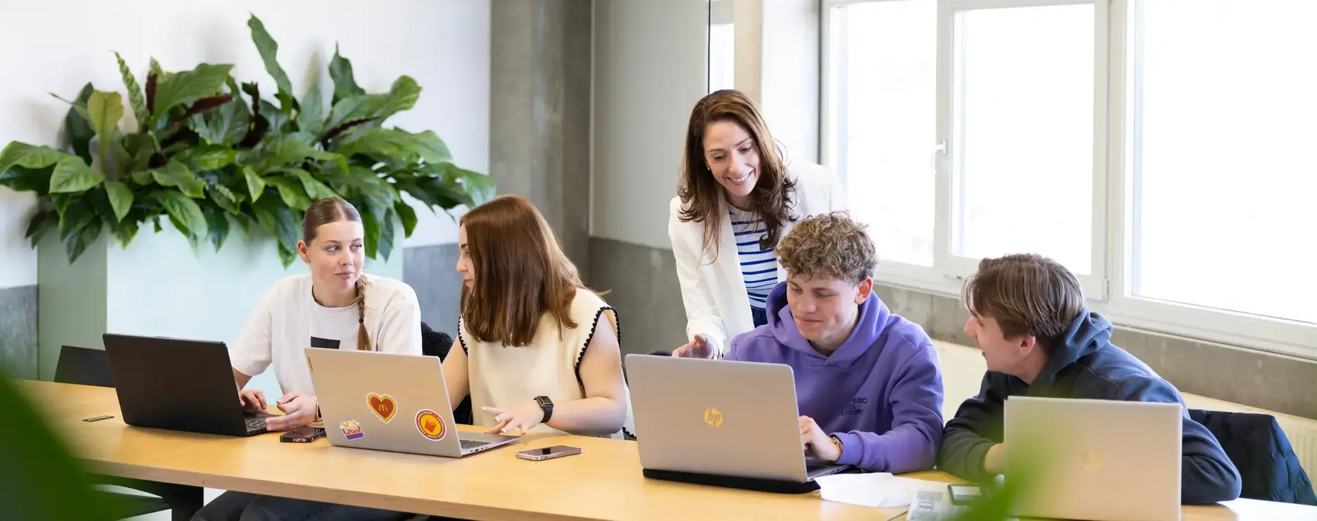 Vier studenten aan een lange tafel met geopende laptops en een docent staat achter hen en legt iets uit.