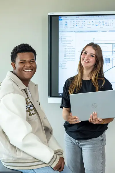 Twee studenten in klaslokaal voor digibord waarop een bouwtekening te zien is. Een student heeft een laptop in haar hand.