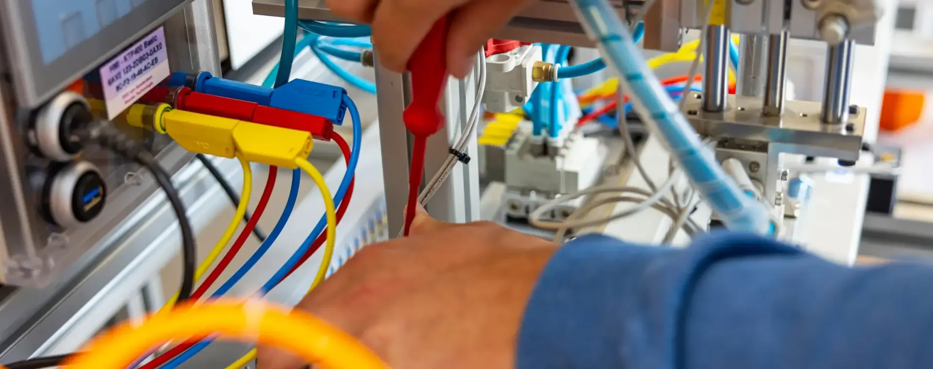 Close-up van handen die bedrading aansluiten op een besturingspaneel met gekleurde kabels en een schroevendraaier, binnen een technische installatie.