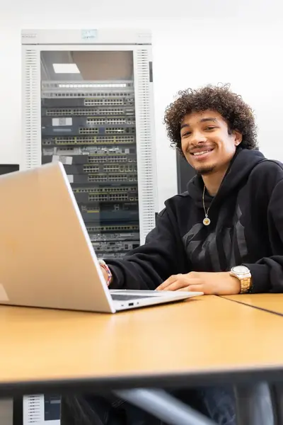 Student werkt op een laptop in een ICT-lokaal met netwerkapparatuur op de achtergrond.
