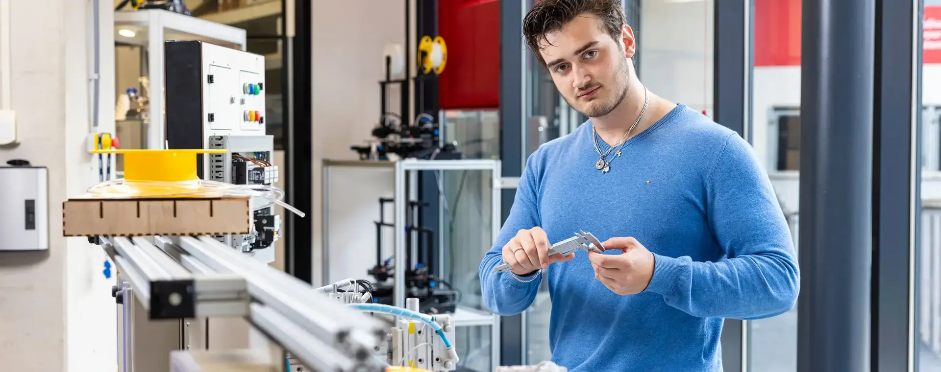 Een jongeman in een blauwe trui staat geconcentreerd bij een technisch werkstation en houdt een schuifmaat vast. Hij is bezig met het nauwkeurig meten van een onderdeel in een techniekomgeving.