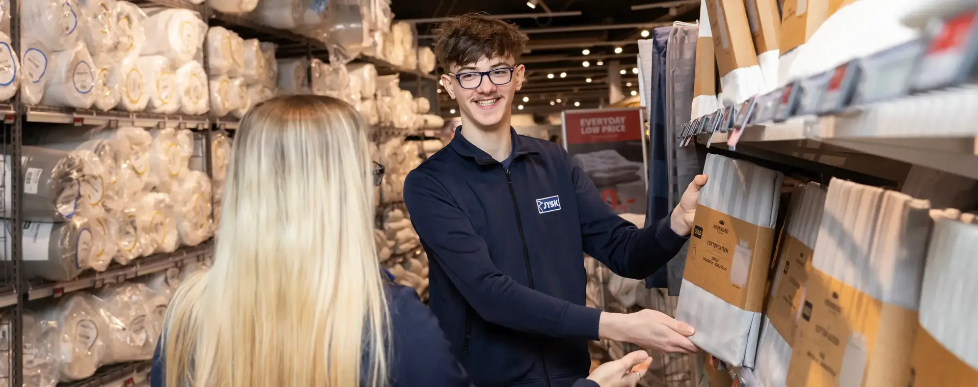 Twee medewerkers staan samen in een winkelgang met bedtextiel. De man wijst glimlachend naar een product.
