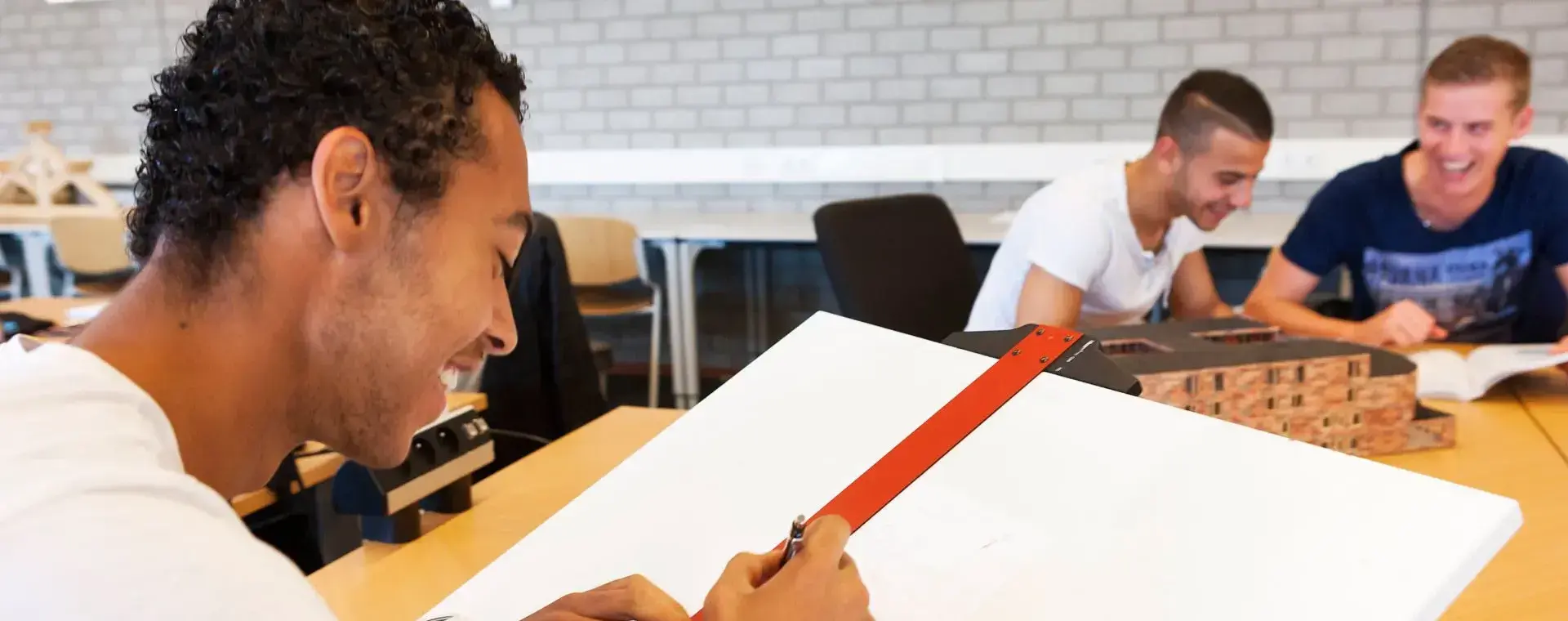 Student tekent op een tekentafel met een tekenhaak. Op de achtergrond werken twee studenten.