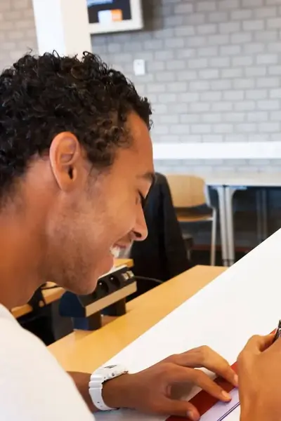 Student tekent op een tekentafel met een tekenhaak. Op de achtergrond werken twee studenten.