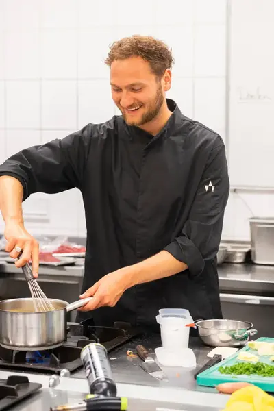 Een jonge kok in zwarte koksbuis staat lachend in een professionele keuken en klopt een mengsel in een pan op het fornuis met een garde.